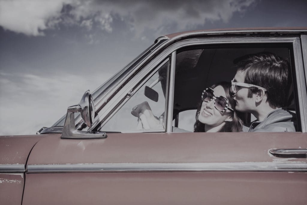 A young man kisses his girlfriend wearing white sunglasses, visible through the driver window of their classic 1960s car.  Vintage styling and colors.  Wide and low angle to emphasis the blue sky and clouds.  Horizontal with copy space.